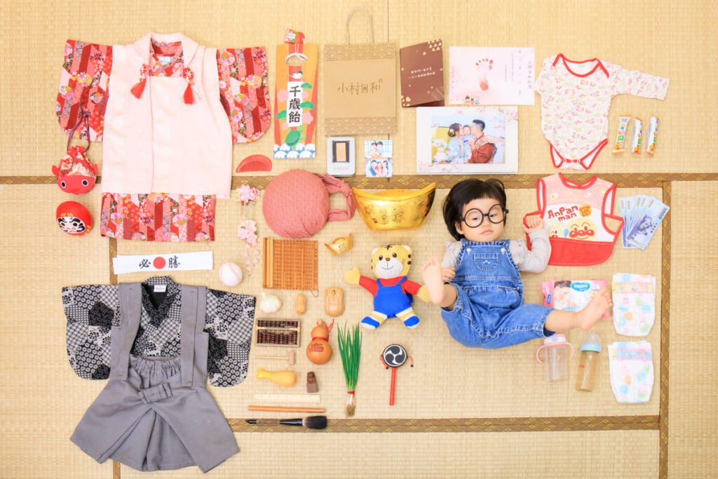 寶寶抓周儀式平拍照，嬰兒躺在榻榻米上，周圍擺放抓周道具、傳統服飾、書籍與紀念物，記錄寶寶一歲的重要成長時刻