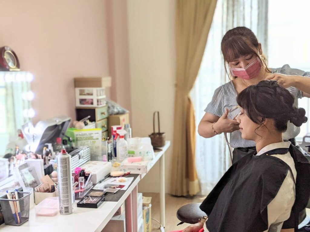 抓周攝影前造型準備畫面，造型師為媽媽整理髮型，梳妝台上擺放化妝與造型工具，記錄抓周活動的幕後時刻
