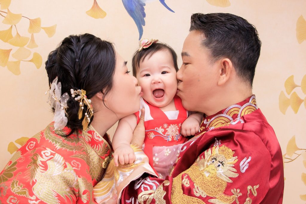 寶寶穿著傳統抓周服飾,開心地被爸媽親吻,溫馨家庭合照