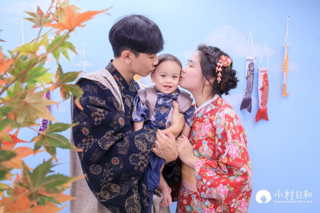 父母身穿日式和服，溫馨地親吻寶寶的額頭，背景有鯉魚旗裝飾，象徵祝福孩子健康成長的美好寓意。