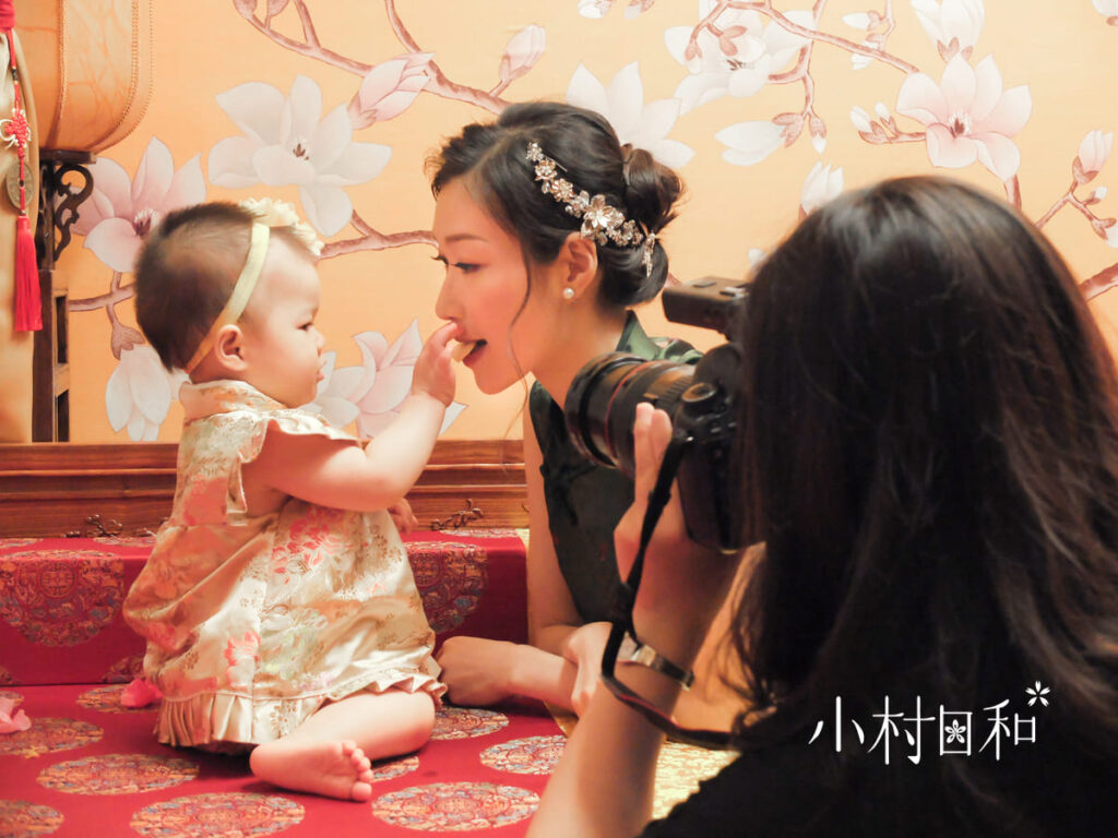 專業攝影師正在抓拍一位寶寶和母親的互動畫面,背景為中式抓周場景,母親穿著精緻的傳統服飾。