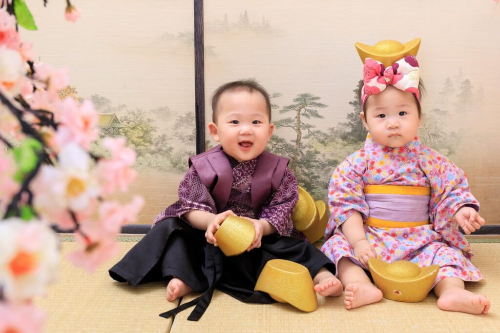 兩位寶寶穿著日式和服,手拿金元寶,坐在日式屏風前,象徵財富與幸福的抓周攝影風格。