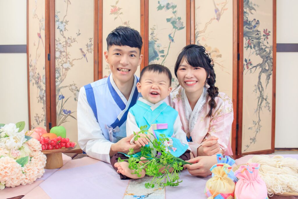 身穿韓服的寶寶與父母坐在傳統韓式布置場景中，手拿水芹象徵健康與長壽，溫馨幸福的抓周儀式。
