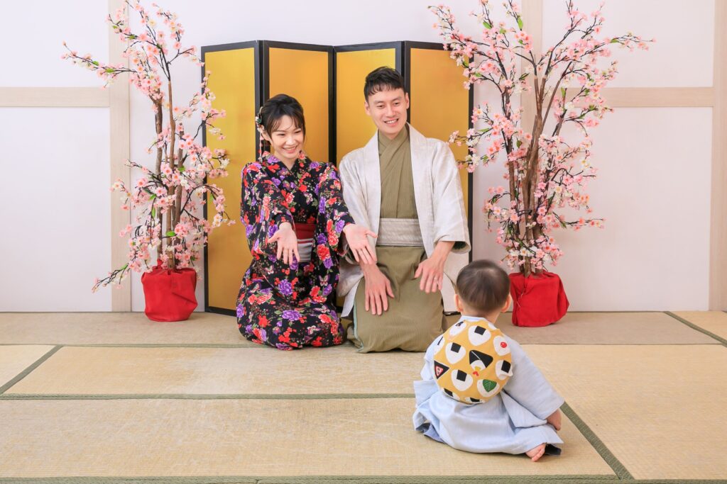 穿著日式和服的父母與寶寶參與傳統抓周儀式,寶寶背負象徵祝福的「一升餅」,背景為櫻花點綴和金色屏風,呈現溫馨的日式家庭氛圍。