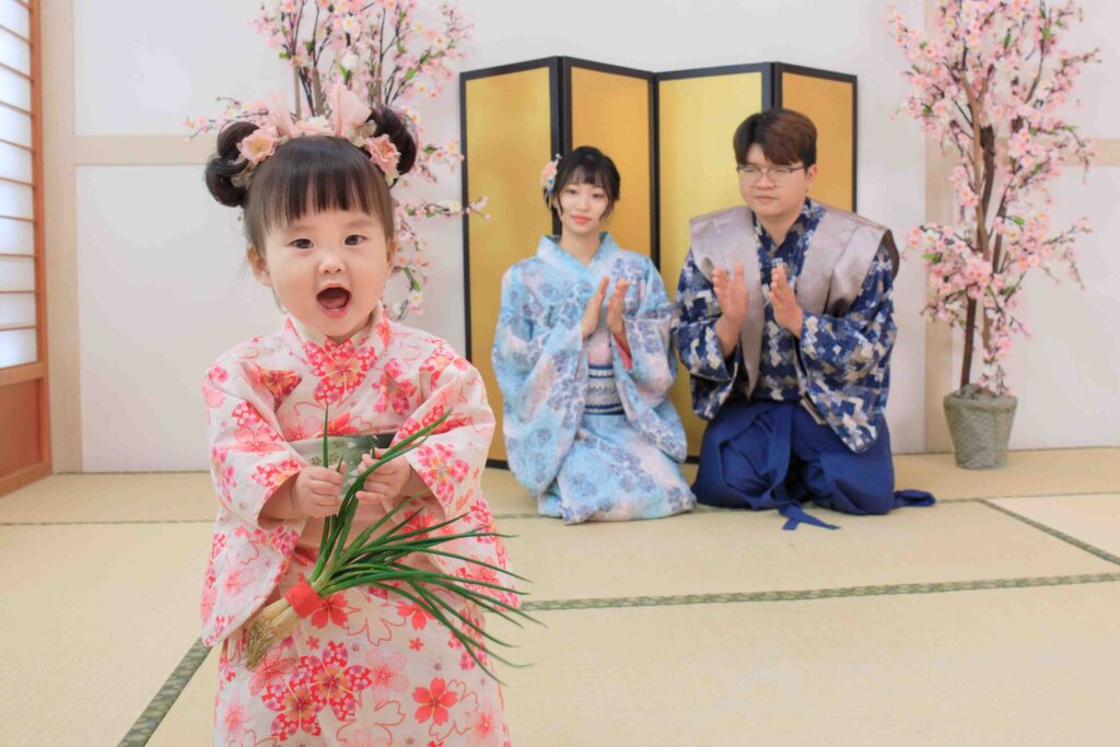 穿著粉色和服的小女孩開心地手持青蔥，背後的家人穿著和服坐在榻榻米上鼓掌，展現日式抓周儀式的傳統氛圍。