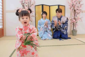 穿著粉色和服的小女孩開心地手持青蔥,背後的家人穿著和服坐在榻榻米上鼓掌,展現日式抓周儀式的傳統氛圍。