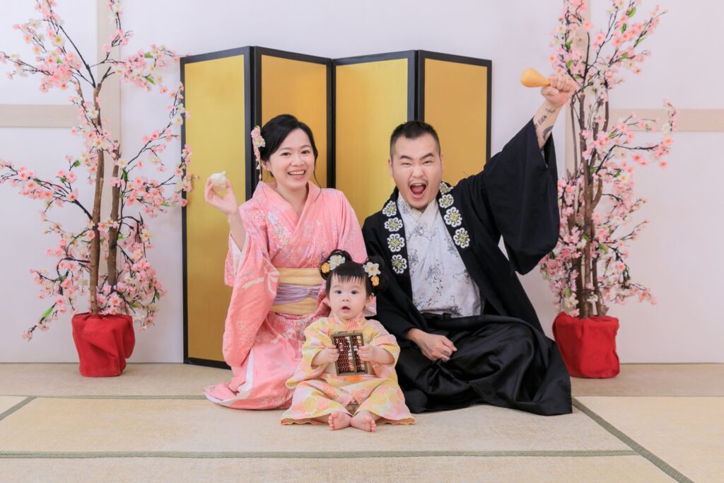 日式家庭抓周儀式場景,父母身著日式和服,陪伴寶寶參與抓周活動,背景為櫻花點綴與金色屏風,捕捉溫馨愉快的家庭時光。