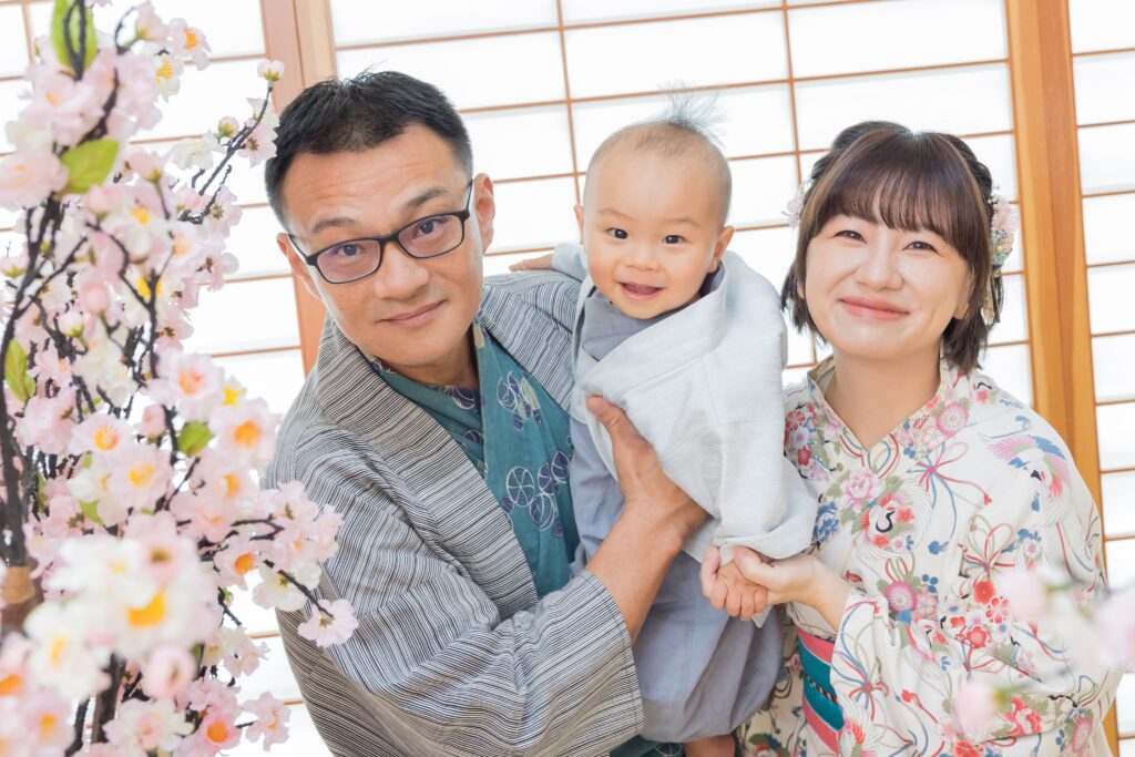 穿著和服的寶寶被父母抱著，父母臉上洋溢著笑容，背景為木質窗戶與櫻花裝飾