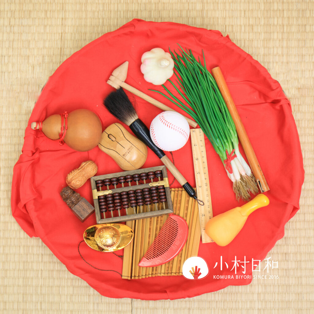 抓周物品全圖展示,包含毛筆、算盤、蔥、蒜、元寶、竹笛等象徵多種未來可能性的道具,擺放在紅色布上,充滿傳統儀式感與文化意義。