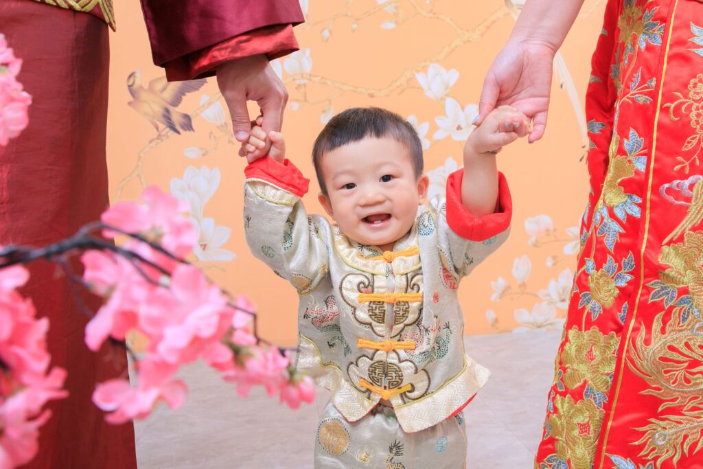 穿著中式傳統服飾的寶寶，在家長的陪伴下開心舉起雙手，背景點綴著桃花與古風元素，象徵幸福與吉祥的抓周儀式。