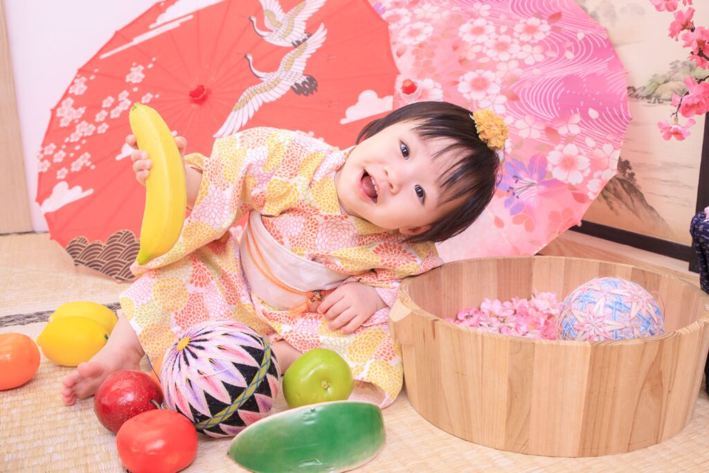 穿著日式傳統服飾的寶寶手握香蕉道具,坐在抓周場景中,背景有鮮豔的紅色和粉色和傘,象徵抓周活動的歡樂氣氛。
