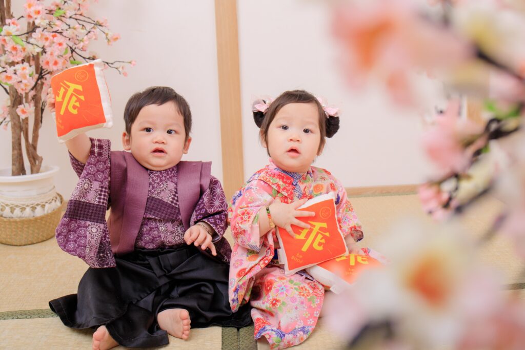 一對可愛的雙胞胎寶寶穿著日式和服，手拿紅色福氣袋，展現可愛模樣，日式抓周儀式中的吉祥畫面。