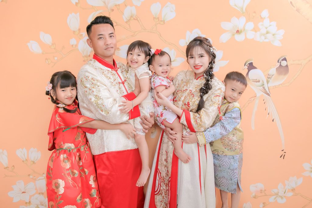 一家六口穿著華麗中式禮服，在橘粉花鳥背景前拍攝全家福，展現熱鬧團圓氣氛。