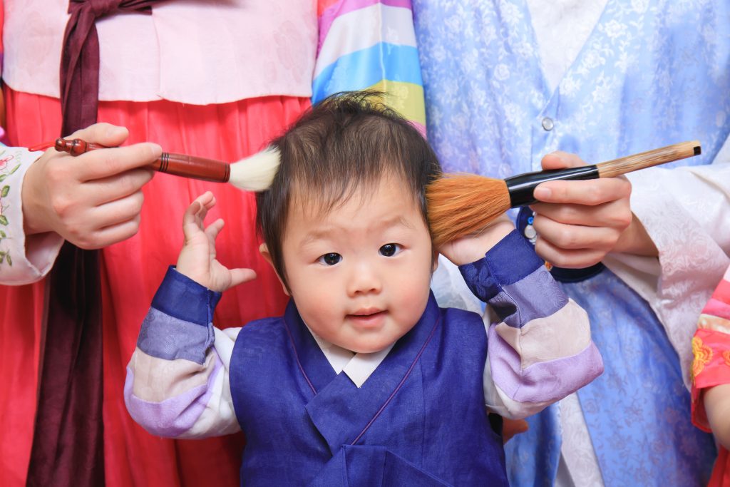 穿著韓服的寶寶參加抓周儀式,家人以毛筆輕掃臉頰,象徵智慧與祝福的溫馨畫面。