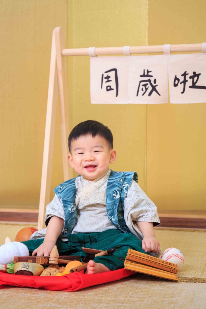 穿著綠色日式傳統武士服的一歲寶寶，開心地坐在抓周物中，進行抓周儀式