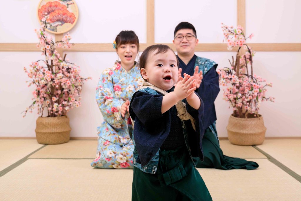抓周儀式現場,滿周歲寶寶穿著和服開心拍手,父母在後方溫馨陪伴紀錄成長時刻。