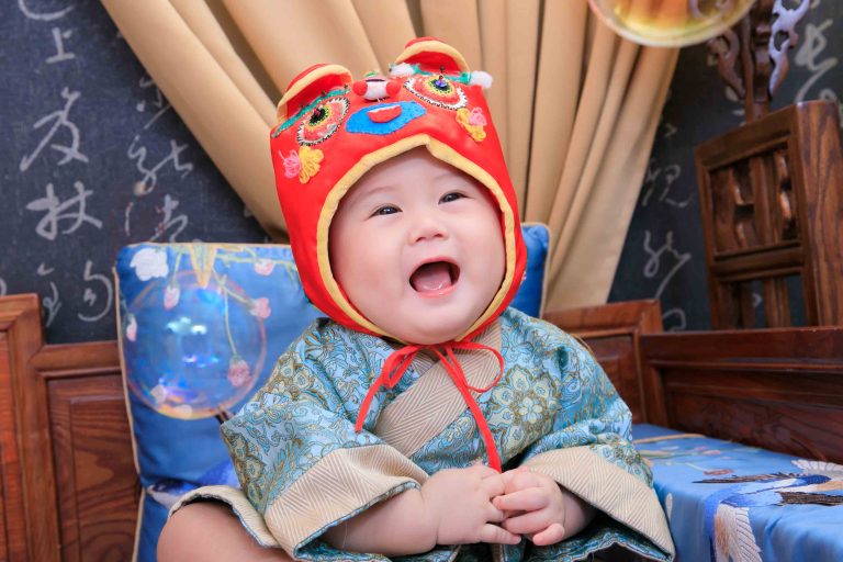 寶寶穿著中式造型服裝，戴紅色虎帽穿傳統服裝開心大笑的一歲紀念畫面