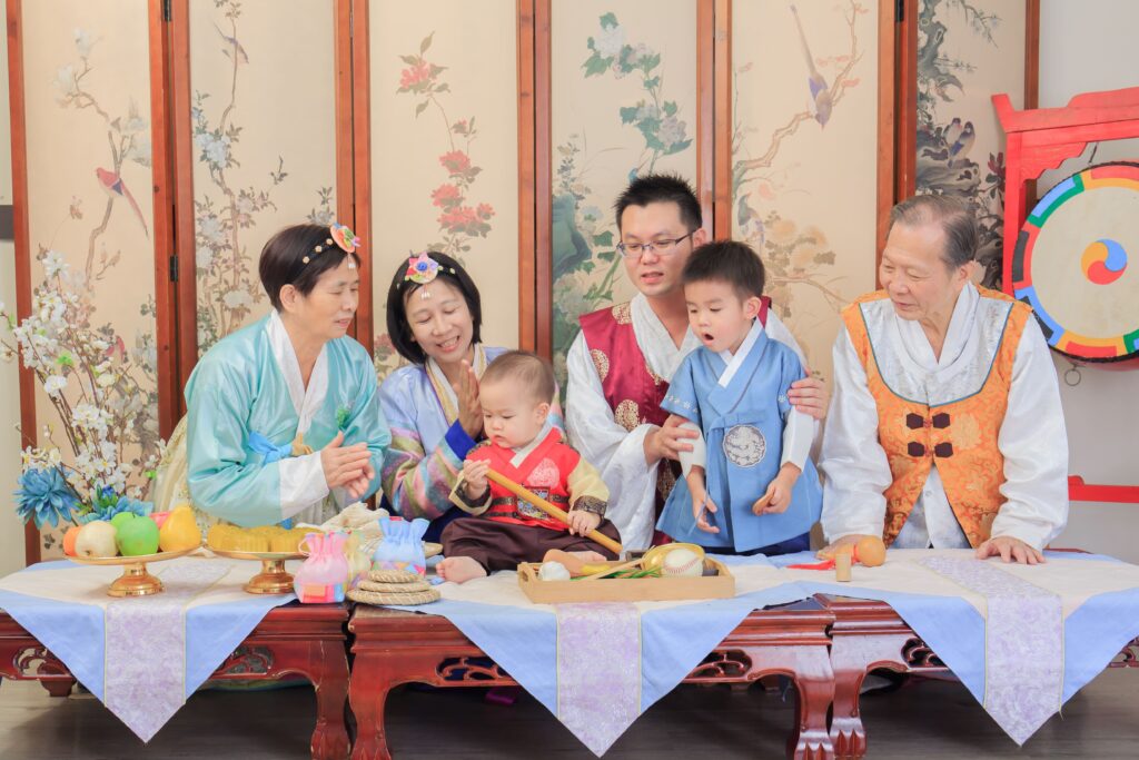 韓式抓周儀式場景，家人身穿傳統韓服，圍繞著穿紅色韓服的寶寶，桌上擺滿象徵意義的抓周物品如棒球、算盤和葫蘆，背景為韓式屏風與傳統裝飾，呈現溫馨歡樂的家庭氛圍與文化儀式感。