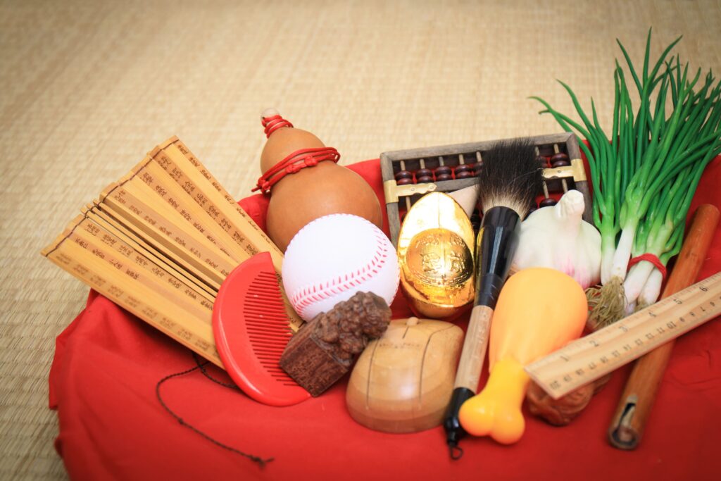 抓周物品展示圖，包含毛筆、金元寶、算盤、籌尺、扇子、紅色梳子、棒球、青蔥、大蒜、葫蘆等，象徵孩子未來不同職業或性格發展的傳統物品，呈現抓周儀式的文化意涵與趣味。