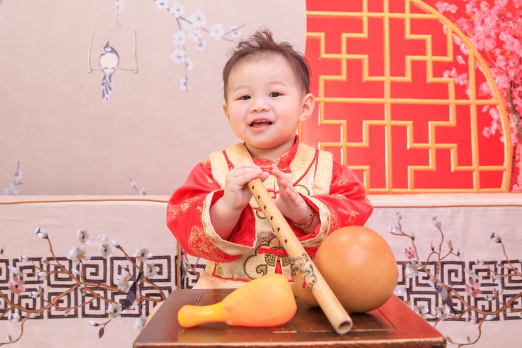 穿著中式紅色傳統服飾的寶寶笑容燦爛，手持象徵抓周儀式的竹笛，桌上擺放葫蘆與玩具，背景為梅花裝飾與中式紅色格紋設計，營造喜慶與溫馨的傳統文化氛圍。