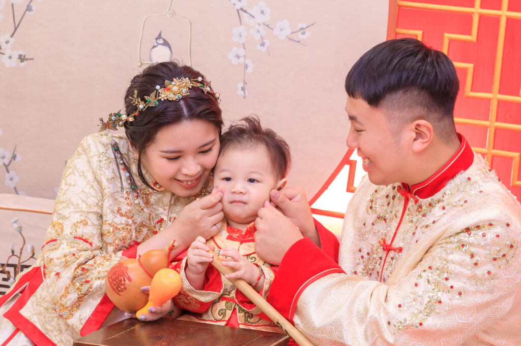 穿著中式傳統服飾的一家三口參與抓周儀式，父母笑著親近寶寶，手持象徵意義的抓周物品如葫蘆與橘子，背景為紅色喜慶裝飾與梅花圖案，展現幸福家庭氛圍與傳統文化儀式的溫馨畫面。