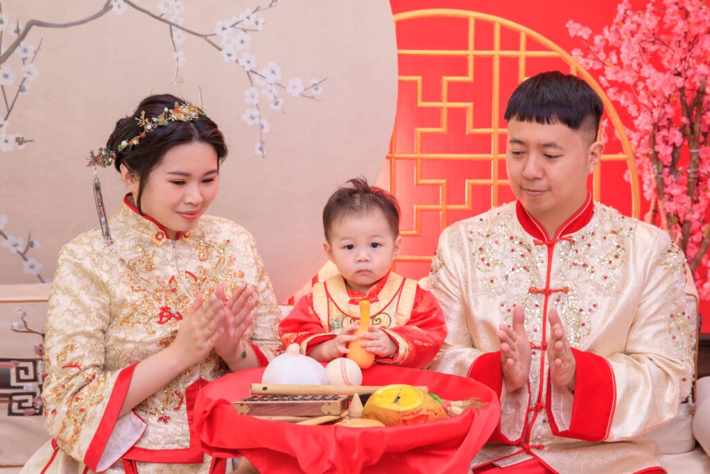 穿著華麗中式禮服的一家三口參與抓周儀式，寶寶坐在正中間，手持抓周物品，桌上擺滿象徵未來的傳統道具，背景為紅色中式裝飾與梅花圖案，展現喜慶與家庭溫馨的抓周文化場景。