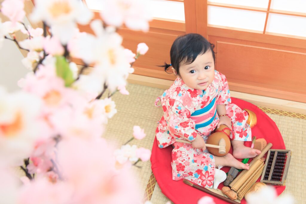 身穿日式花紋和服的可愛寶寶坐在包著紅布的竹籃上，面前擺滿象徵抓周意義的物品如算盤、毛筆和葫蘆，背景為櫻花與日式拉門，呈現傳統與童趣結合的溫馨抓周場景。