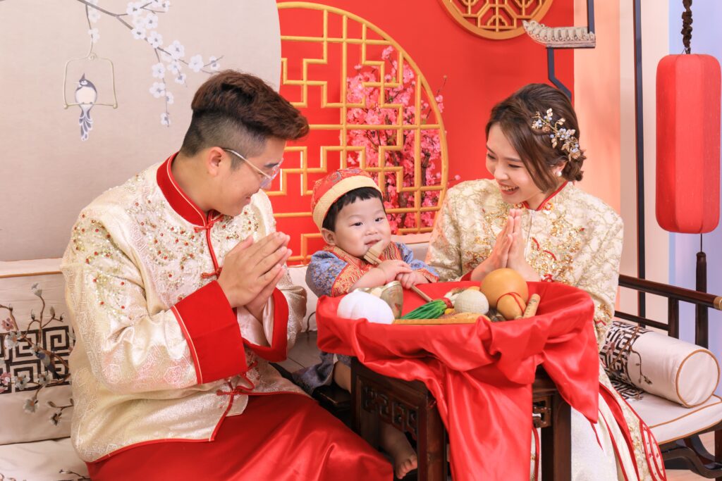 身穿華麗中式禮服的一家三口參與抓周儀式，寶寶坐在紅色抓周布前，面前擺滿抓周物品，背景為喜慶的中式裝飾，展現溫馨與祝福的傳統家庭儀式。