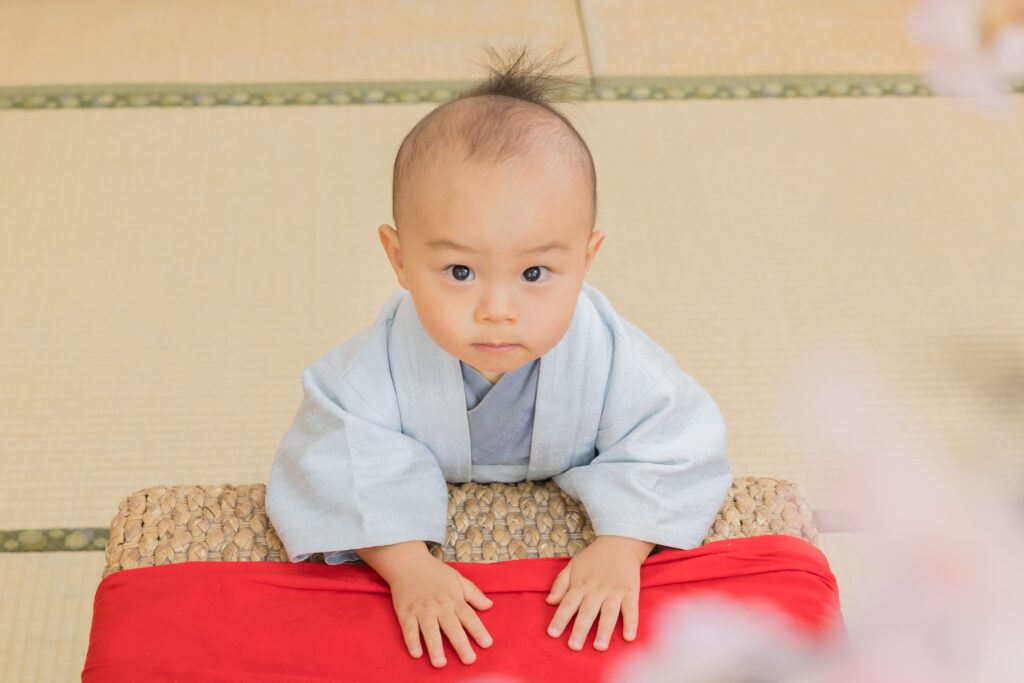 穿著藍色和服的寶寶，專注地坐在紅色布上，表情可愛，背景為傳統和式榻榻米。