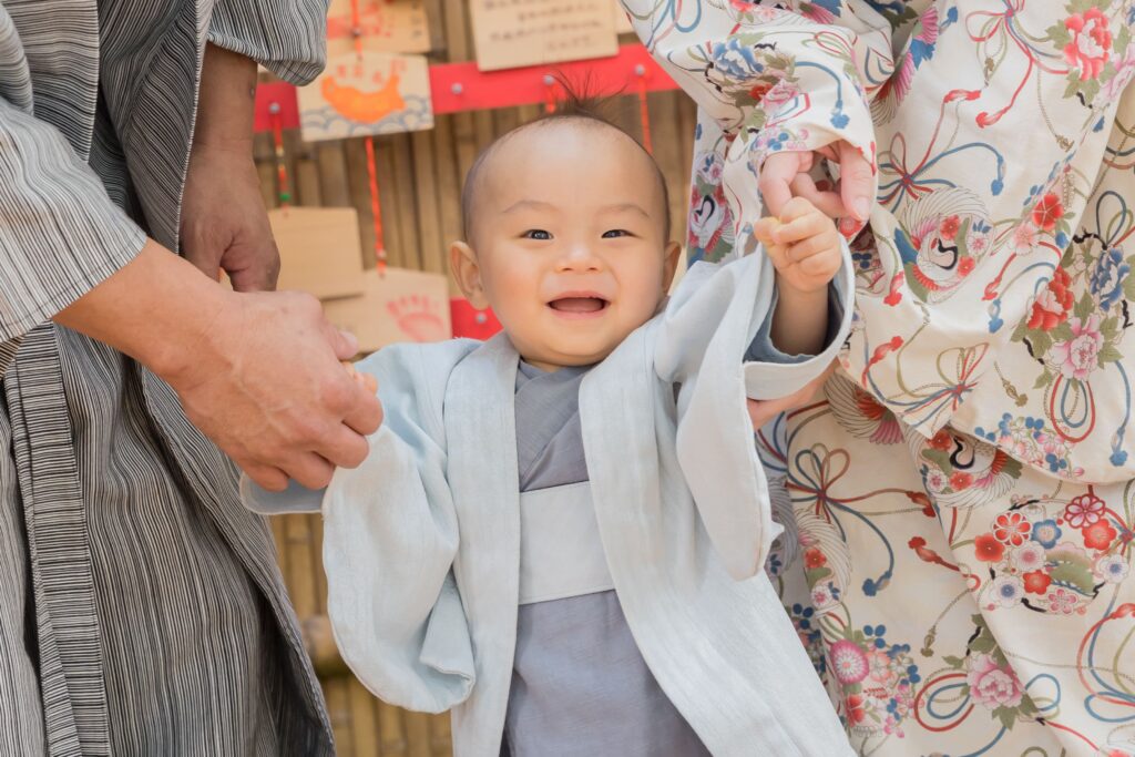 寶寶穿著傳統藍色和服，在父母的牽引下站立，開心大笑，背景為日本繪馬和紅色裝飾。