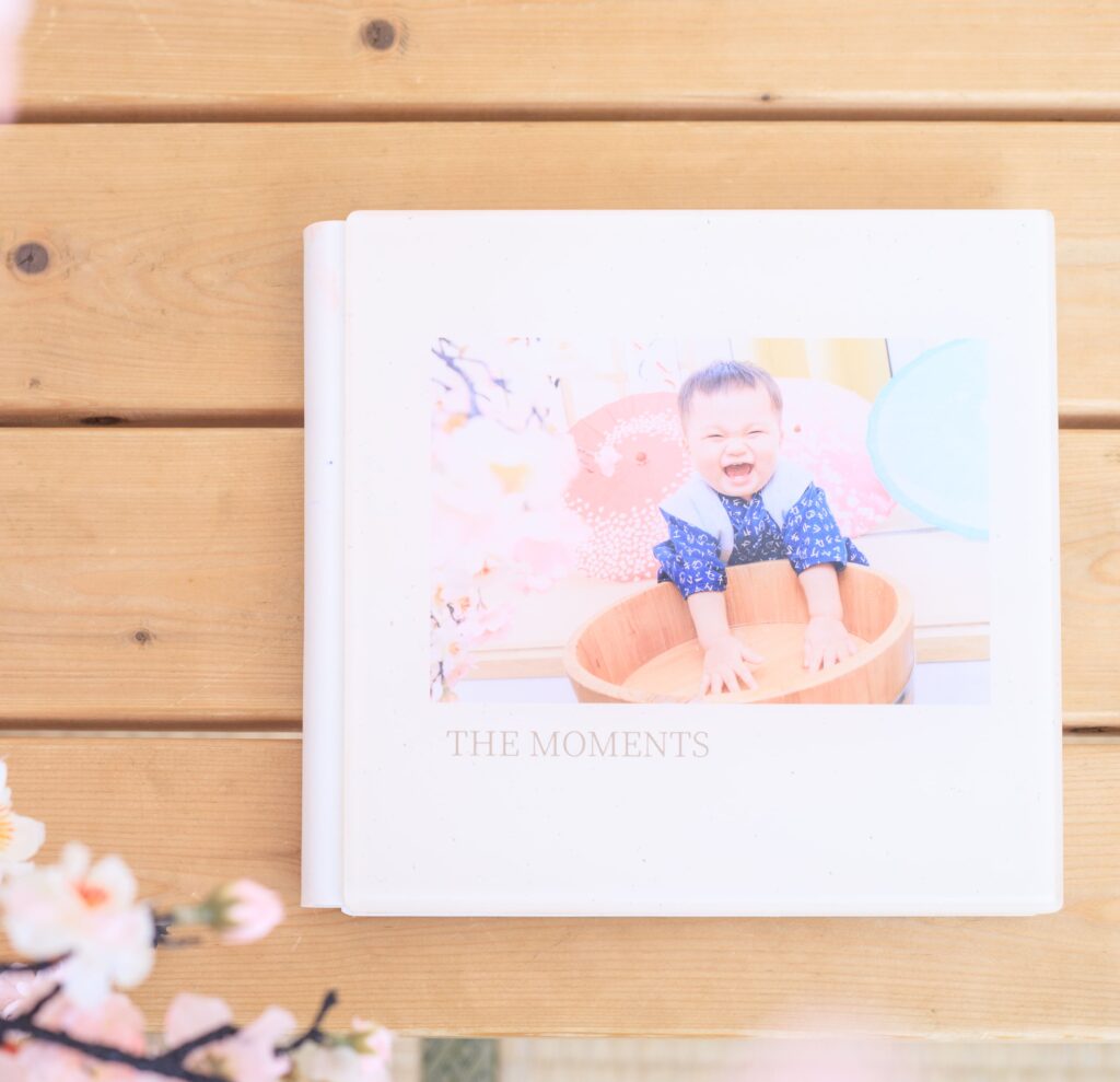 一本放置在木桌上的抓周攝影相冊，封面展示了一位笑容燦爛的寶寶，背景有櫻花點綴。