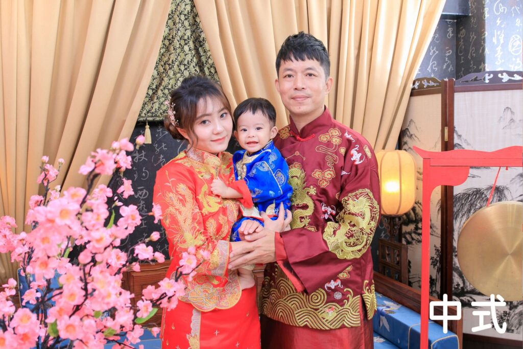一家三口穿著傳統中式禮服，在抓周攝影棚中合影，背景為古風屏風與紅色佈景，展現濃厚的東方文化氛圍。