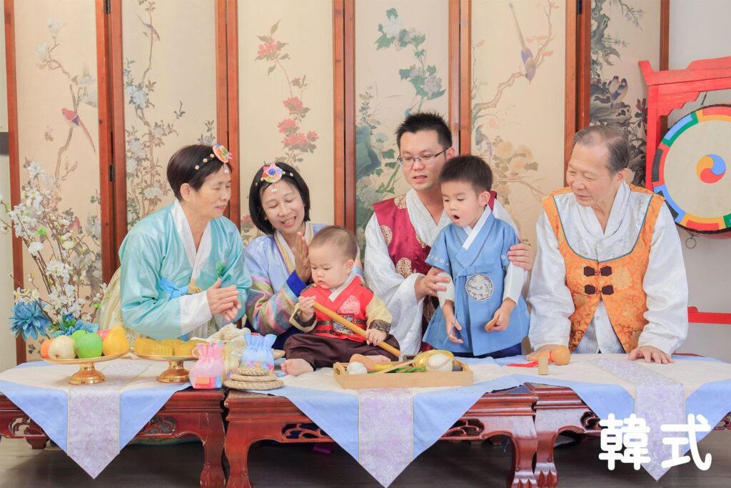 全家人穿著韓服，寶寶在抓周儀式中挑選道具，桌上擺滿傳統韓式抓周用品，呈現韓式風格的攝影場景。