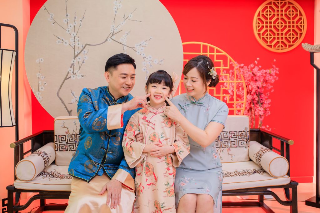 一家三口穿著傳統中式服飾，在紅色背景與梅花裝飾前拍攝幸福全家福照
