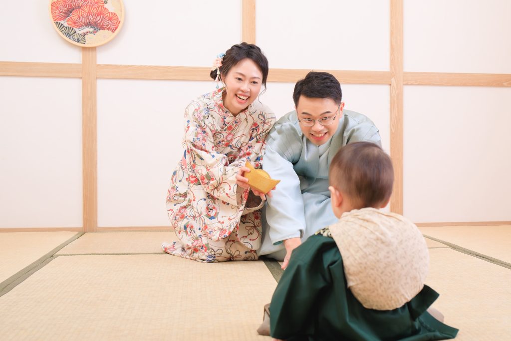 爸媽穿著和服,手拿著金元寶正鼓勵寶寶抓取,臉上表情興奮,象徵吉祥祝福的抓周活動場景。