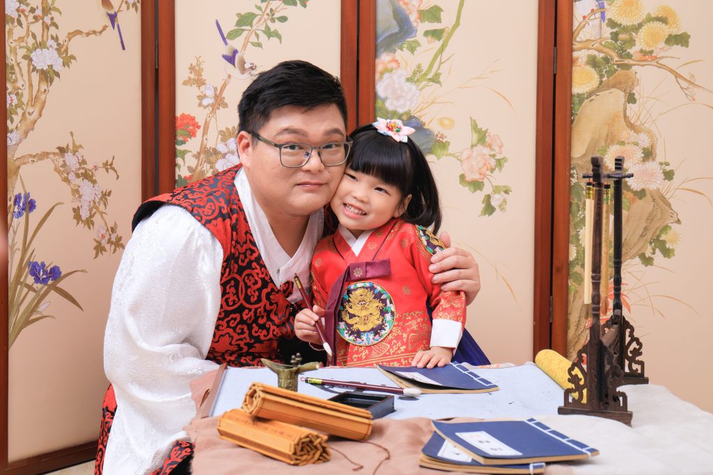 父女兩人穿著韓國傳統服飾,坐在書桌前,爸爸摟著女兒溫馨合影,展現深厚的親子情感。