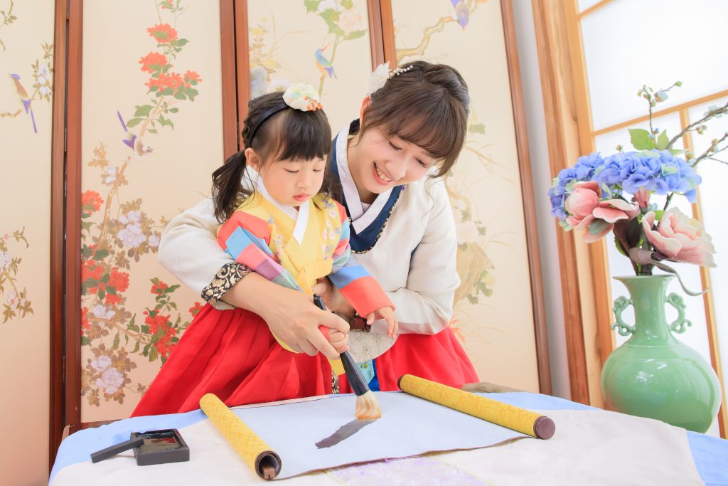 溫馨母女穿著韓服在傳統花鳥屏風前練習毛筆書法,媽媽手把手教小女孩揮毫,桌上擺有宣紙、墨盒與卷軸,旁邊放著插有藍色繡球花與粉色花朵的花瓶,呈現親子文化體驗與傳統藝術學習氛圍。