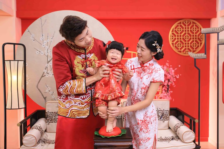 一家三口穿著紅色傳統服飾慶祝節日，父母微笑著扶著穿紅色洋裝的嬰兒站在小凳子上，背景佈置有紅色牆面、花卉與中式裝飾。