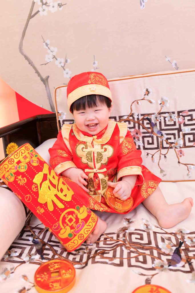 穿著紅色傳統服飾的一歲寶寶開心坐在佈景中,手拿鞭炮及金幣