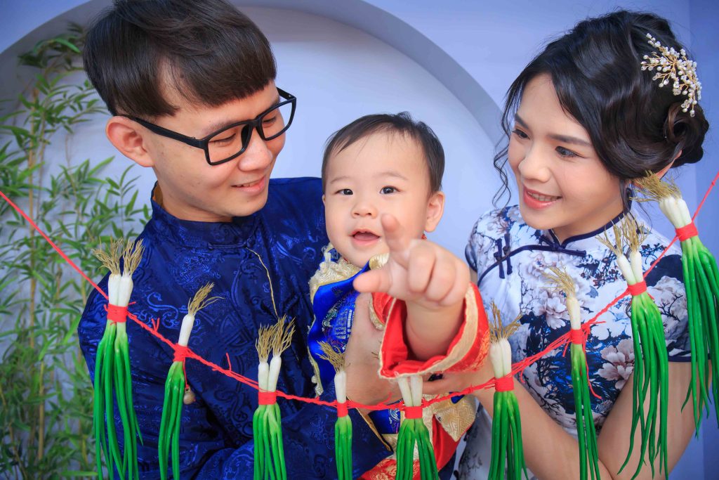 一家三口穿著傳統服飾微笑合影，父親抱著嬰兒，嬰兒伸手指向前方，前景掛著綁有紅線的青蔥裝飾，背景有綠色植物與拱形牆面。