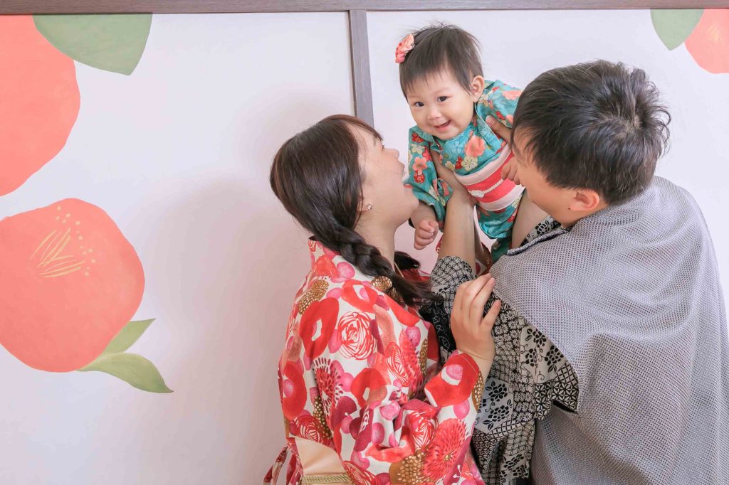 一家三口穿著傳統和服風格服飾互動，父母抱起穿著綠色花紋衣服的嬰兒，嬰兒開心微笑，背景為淡色牆面與花卉圖案裝飾。