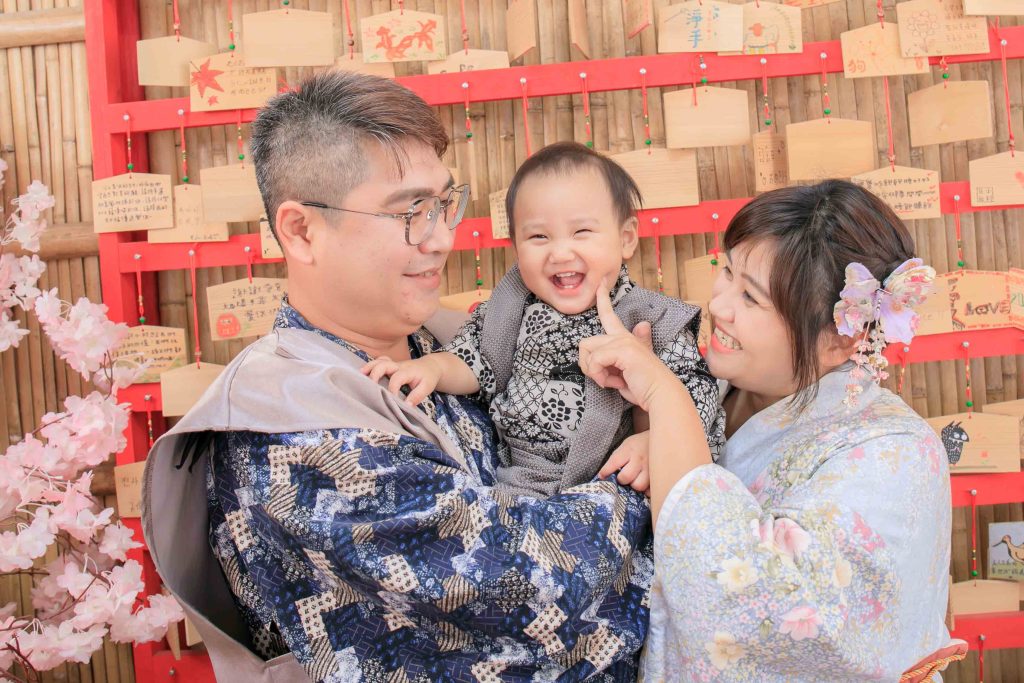 父母穿著傳統服飾抱著微笑寶寶合影，背景為祈福繪馬牆的一歲紀念照片