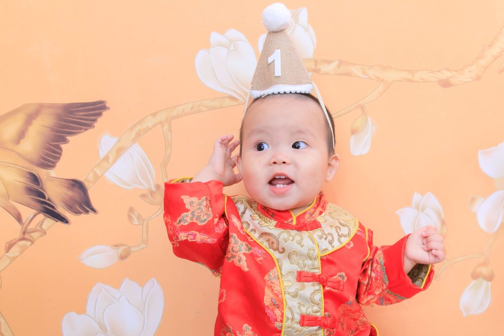 寶寶一歲抓周紀念,小男嬰穿紅色中式服裝戴生日帽,呈現喜氣溫馨畫面