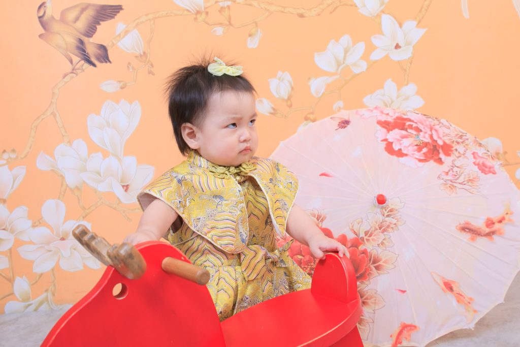 寶寶抓周紀念畫面，小女孩穿中式服裝在花卉與油紙傘場景中互動，呈現典雅氛圍